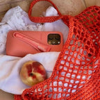 Coral phone case on a white cloth with a peach, next to an orange mesh bag.