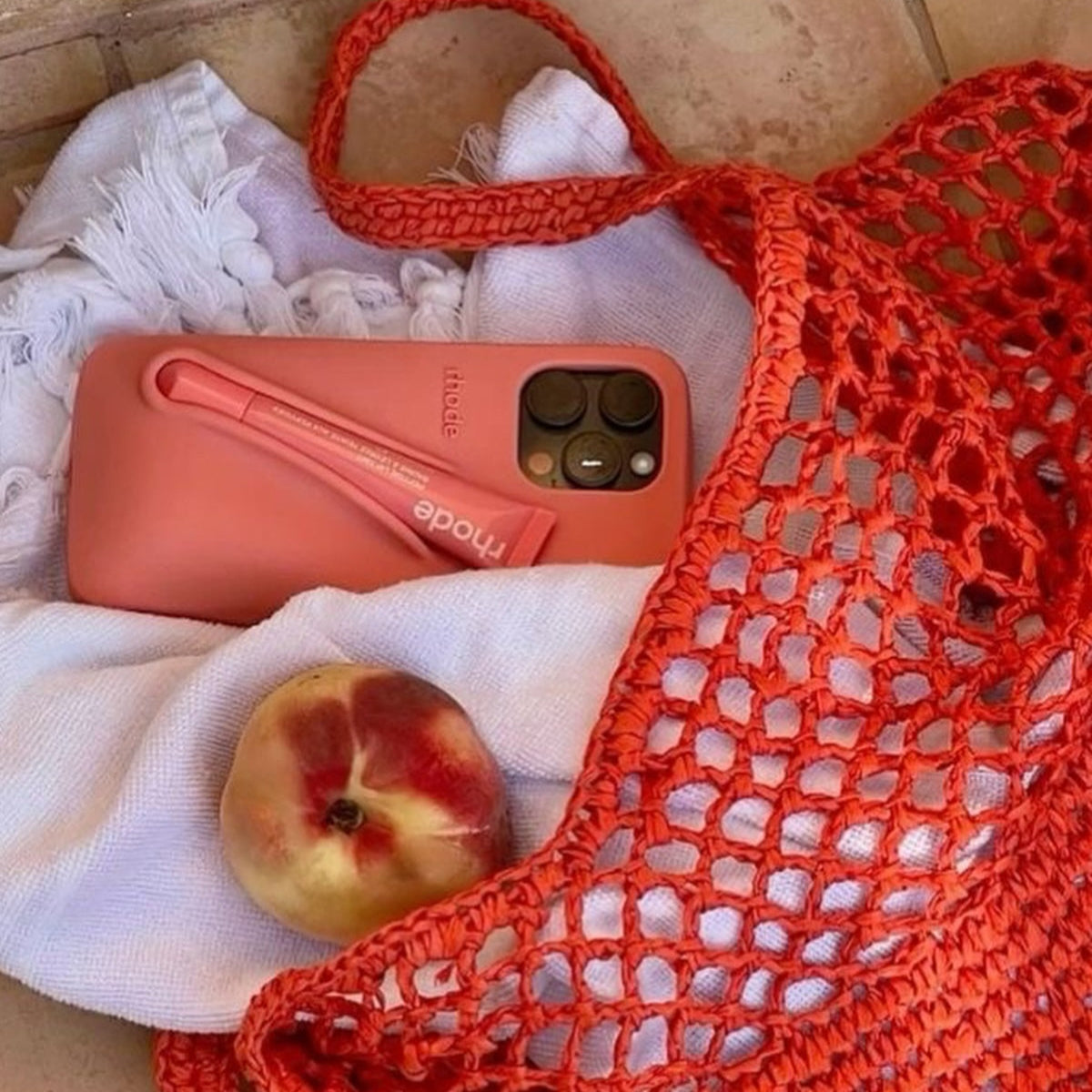 Coral phone case on a white cloth with a peach, next to an orange mesh bag.